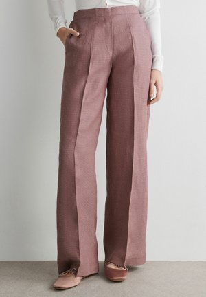 Donna che indossa pantaloni a vita alta a gamba larga color malva abbinati a ballerine coordinate e una camicia bianca con bottoni, in piedi all'interno.