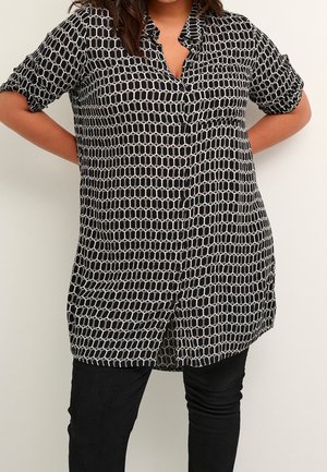 Femme portant une tunique chemise à boutons avec un motif géométrique noir et blanc, manches retroussées, et un pantalon noir. Les mains sont derrière son dos.