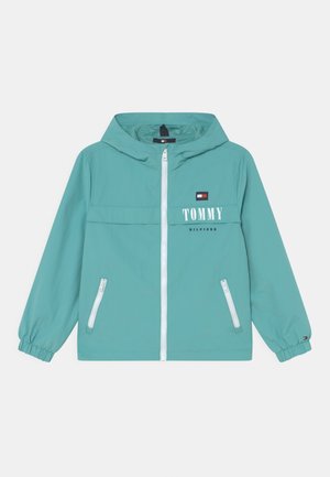 Coupe-vent bleu-vert clair avec un zip à l'avant, une capuche, des poignets élastiques et deux poches latérales zippées. Présente un logo imprimé blanc sur la poitrine.