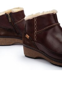 Botas de tobillo de cuero marrón con forro de piel sintética, cremallera lateral y suelas de cuña texturizadas. Presenta costuras contrastantes y una etiqueta de la marca.