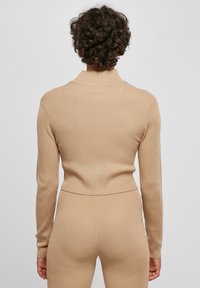 Beige ribbad stickad tröja med hög krage och långa ärmar, åtsittande vid midjan, med en slät yta och minimal detaljering.