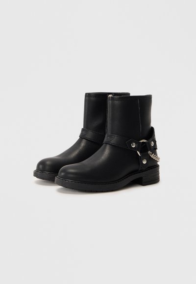 Stivali neri in pelle alla caviglia con suola piatta, cinturino laterale con fibbia e dettaglio in catena. Presentano una texture liscia e un design minimalista.