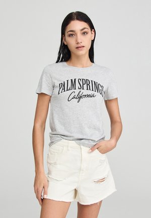 T-shirt grigia in cotone con la scritta "PALM SPRINGS California" stampata in nero, abbinata a pantaloni corti in denim bianchi con effetto usurato e orlo sfilacciato.