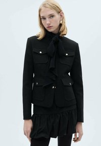 Sacou negru croit, cu detalii aurii, două buzunare în față, o cravată la gât și o margine frotiată. Fabricat dintr-un material structurat cu o textură netedă.