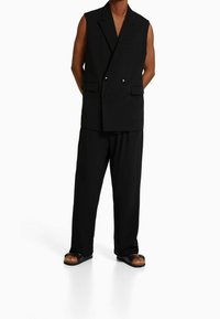 Svart ärmlös dubbelknäppt väst med slag, i kombination med lösa svarta byxor. Bärs med bruna sandaler med en tjock svart sula.