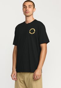 T-shirt noir en coton à manches courtes avec un graphique circulaire jaune comportant le texte "VOLCOM STONE" et "TIME TO TRAVEL" sur le devant.