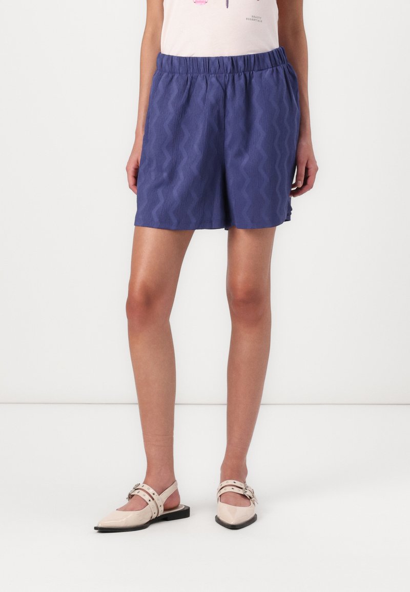 Paarse shorts met een zigzagtextuur, voorzien van een elastische tailleband. Gecombineerd met beige platte schoenen. Achtergrond is eenvoudig wit.