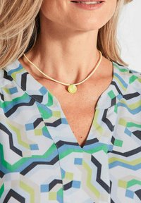 Beige Choker-Halskette mit einem runden gelben Anhänger, getragen mit einer mehrfarbigen Bluse im geometrischen Muster, die grüne, blaue und schwarze Akzente aufweist.