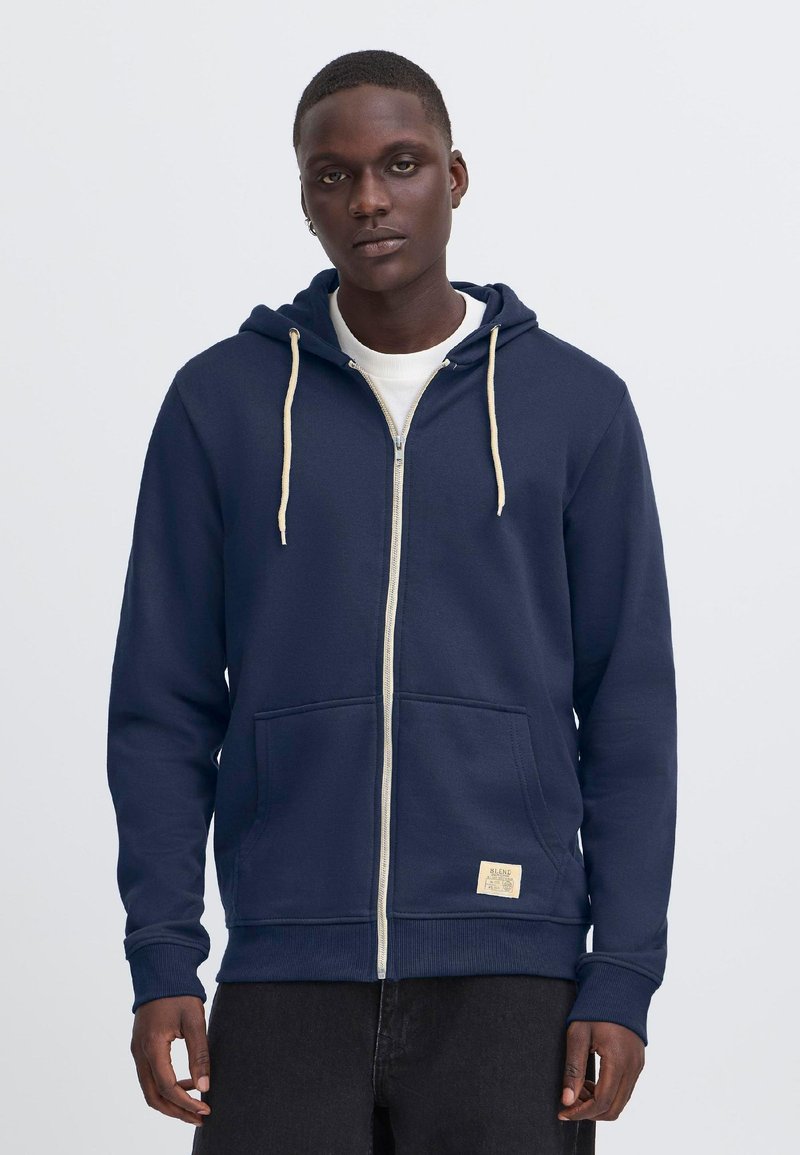 Männlich, trägt einen marineblauen Hoodie mit Reißverschluss, cremefarbenen Kordeln und einem Aufnäher, steht vor einem schlichten hellgrauen Hintergrund.