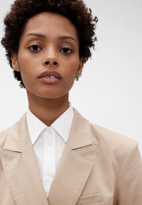 Bež sako s urezanim reverima, jednim džepom i teksturiranim materijalom u kombinaciji s bijelom košuljom s ovratnikom i kopčanjem na gumbe.