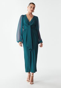 Türkisfarbenes, strukturiertes Kleid mit tiefem V-Ausschnitt, langen transparenten Ärmeln und einem Knoten vorne. Kombiniert mit weißen Riemchen-Heels.