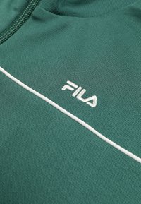 Green textured fabric with white FILA logo embroidery and a thin white horizontal stripe, featuring a partially visible zipper at the top.