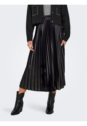 Frau trägt eine schwarze Jacke mit Nieten, einen metallisch plissierten Midirock und schwarze Stiefeletten mit Absatz vor hellgrauem Hintergrund.