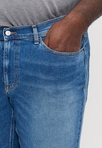 Blå denimjeans med tapered passform, som har en framficka, kontrasterande sömmar och knäppning. Tyget har en slät yta.