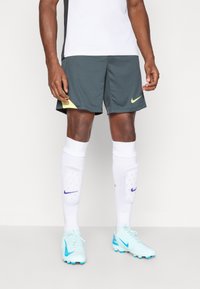Calças de desporto cinzentas com detalhes amarelos, meias brancas até aos joelhos e botas de futebol azul claro. Apresenta logotipos da Nike nas calças e nas meias.