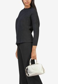Zwarte gebreide trui met een hoge halslijn en verlaagde schouders, gecombineerd met een zwarte broek. Houdt een witte handtas van patentenleer met een krokodillenstructuur vast.