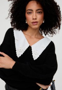 Schwarzer Pullover mit einem großen weißen, bestickten Kragen, der florale Muster und geschwungene Kanten aufweist; Model hat lockiges Haar und hält einen Griff.