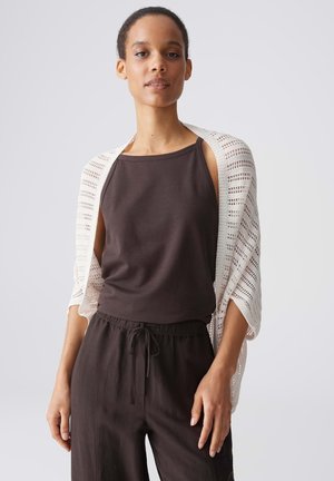 Donna che indossa un top marrone scuro senza maniche e pantaloni con uno scialle bianco a maglia aperta drappeggiato sulle spalle, in piedi davanti a uno sfondo chiaro e semplice.