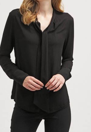 Kobieta ubrana w czarną bluzkę z długim rękawem z luźnym wiązaniem przy kołnierzu, w połączeniu z czarnymi spodniami na jednolitym tle.