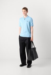 Camisola polo azul claro com fecho, logótipo preto. Combinada com calças pretas largas e uma grande mala preta. Texturas suaves.