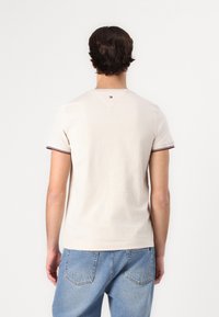 Beige bomull T-skjorte med korte ermer, har mørkerøde og marineblå detaljer på kragen og ermene, og en liten logo bak.