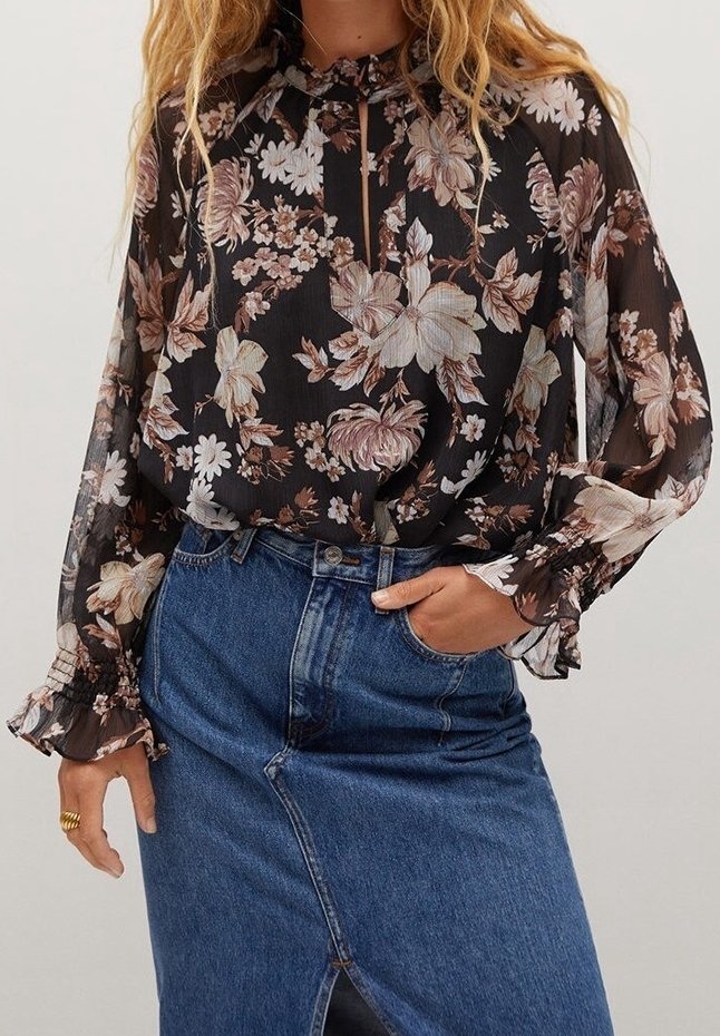Femme portant un chemisier noir transparent à motif floral avec des poignets à volants, rentré dans une jupe en jean bleue taille haute avec une fente à l'avant.