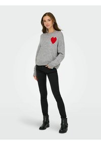 Maglione grigio con un accento a forma di cuore rosso, tessuto morbido, vestibilità ampia. Abbinato a jeans skinny neri e stivaletti neri alla caviglia. Design minimalista.