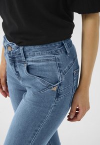 Světle modré rifle s vysokým pasem, se pěti kapsami, stříbrným zapínáním na knoflík a trojúhelníkovým logem na zadní kapse.