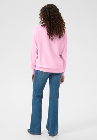 Lichtroze trui met geribde mouwen en zoom, gecombineerd met blauwe wijde jeans, gezien vanaf de achterkant. Krullend haar zichtbaar.
