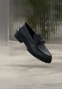 Μαύρο δερμάτινο μοκασίνι με στρογγυλεμένη μύτη, υφή στο εμπρόσθιο μέρος και διακοσμητικό μεταλλικό στοιχείο. Διαθέτει χοντρή ελαστική σόλα για μεγαλύτερη πρόσφυση.