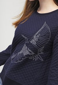 Navy quilted sweatshirt with a textured finish, featuring a white embroidered eagle design on the front, round neckline, and long sleeves.