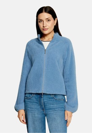 Vrouw draagt een lichtblauw, pluizig jasje met rits en blauwe spijkerbroek, staand tegen een effen witte achtergrond.