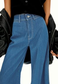 Široke denim hlače v svetlo modri barvi, z visokim pasom, kontrastnim šivom in kovinskim gumbom na sprednji strani.