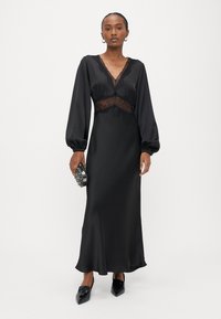 Melna satīna kleita ar garām piedurknēm ar pūkainiem piedurkņu galiem, mežģīņu ieliktni viduklī un plūstošu siluetu. Modele tur rokā rotaslietu somiņu un valkā smailus loaferus.