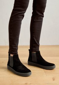 Schwarze Wildleder-Ankle Boots mit strukturierten grauen elastischen seitlichen Einsätzen und einer geriffelten Sohle. Getragen mit dunklen, engen Hosen auf einem Holzboden.