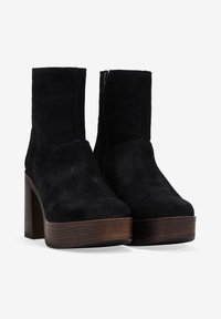 Botas de tobillo de ante negro con tacones altos de bloque y suelas de plataforma de madera marrón, con cierres de cremallera lateral, sobre un fondo blanco.