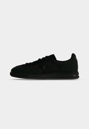 Baskets noires à faible profil avec fermeture à lacets, semelle lisse et logo discret sur le côté et le talon, présentées en vue latérale sur fond uni.