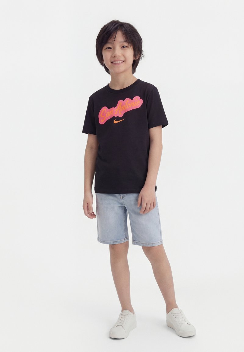 Lachende jongen staand, gekleed in een zwart "San Antonio" Nike T-shirt, lichte denim korte broek en witte sneakers, tegen een effen witte achtergrond.