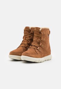 Botas de tobillo de ante marrón con forro de piel de oveja color crema, puntera redonda, suela plana de goma y diseño de encaje con múltiples ojales y botones laterales.