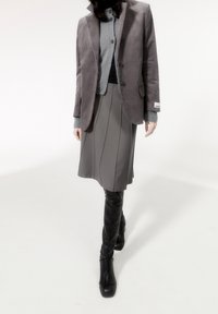 En kvinna i grå kjol, grå stickad tröja, grå blazer och svarta knälånga läderstövlar som går framåt mot en vit bakgrund.