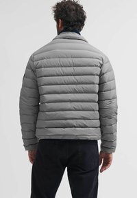 Sivi puffer kaput s horizontalnim šavovima, visokim ovratnikom i rebrastim detaljima. Materijal deluje glatko i lagano, pružajući izolaciju.