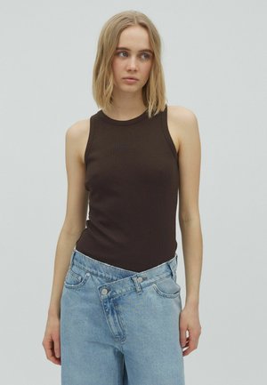Jeune femme aux cheveux blonds mi-longs, portant un haut noir sans manches et un jean bleu clair oversize sur un fond clair uni.