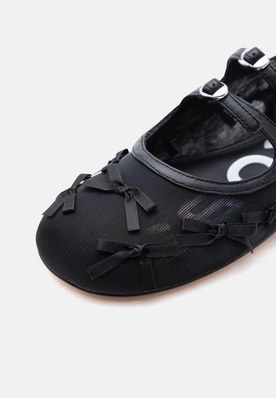 Μαύρη μπαλαρίνα με διαφανές ύφασμα, διακοσμητικά μικρά φιόγκια μπροστά και στα πλάγια, και διπλά ασημένια λουράκια με αγκράφα γύρω από τον αστράγαλο.