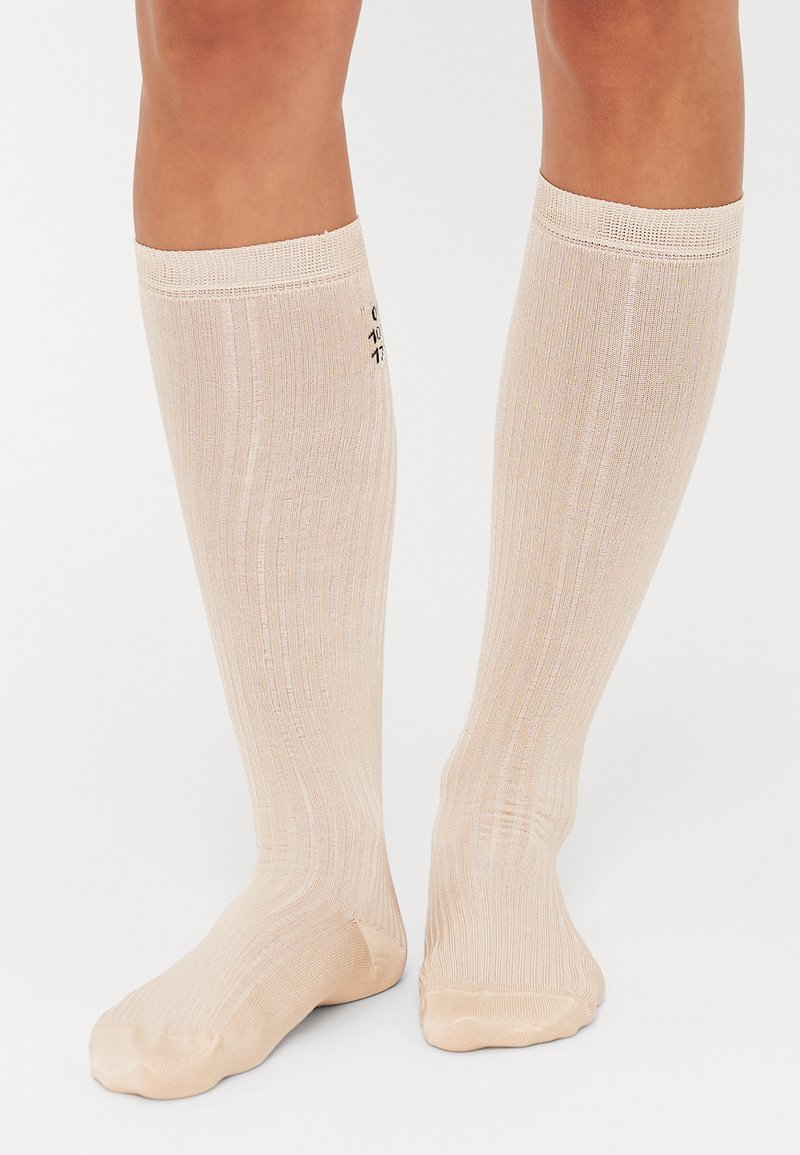 Chaussettes montantes de couleur beige clair avec une texture côtelée, dotées d'un léger éclat et d'un petit logo imprimé sur le côté.