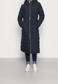 Abrigo largo acolchado en azul marino oscuro con capucha, cremallera frontal y diseño acolchado vertical; combinado con vaqueros ajustados y zapatillas blancas.