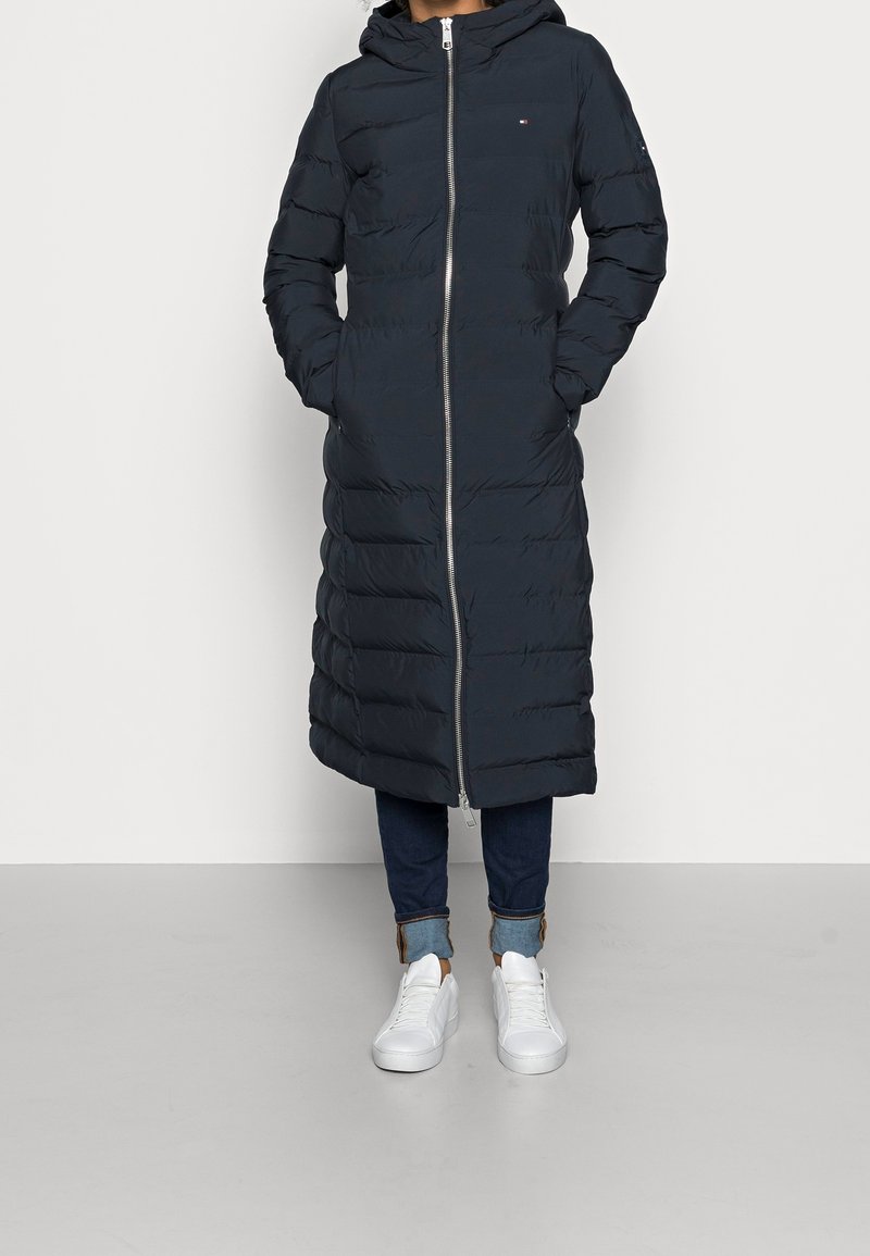 Abrigo largo acolchado en azul marino oscuro con capucha, cremallera frontal y diseño acolchado vertical; combinado con vaqueros ajustados y zapatillas blancas.