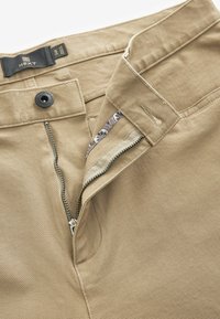 Pantalons kaki avec une fermeture éclair et un bouton. Tissu texturé avec un intérieur de ceinture lisse présentant des détails en motif.