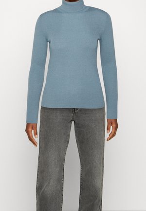 Person trägt einen hellblauen Langarm-Rollkragenpullover und hoch taillierte graue Jeans, steht vor einem weißen Hintergrund.