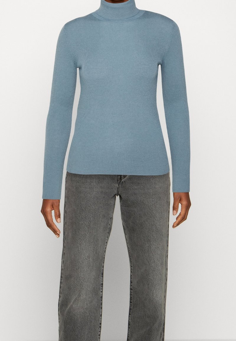 Personne portant un pull à col roulé bleu clair à manches longues et un jean gris taille haute, debout devant un fond blanc.