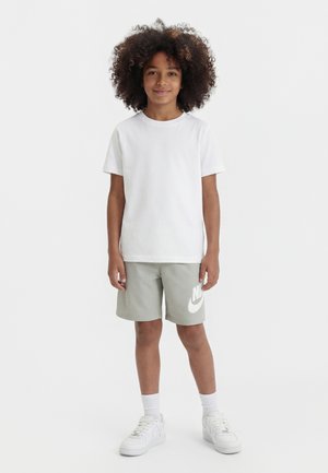 Enfant aux cheveux bouclés portant un t-shirt blanc, un short Nike gris clair, des chaussettes blanches et des baskets blanches, debout devant un fond blanc.
